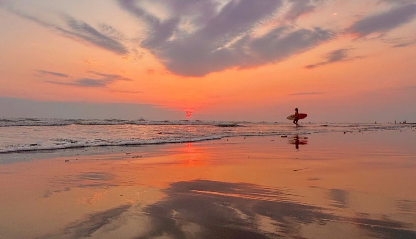 A man holding a surfboard walking on a Costa Rica beach at sunset.